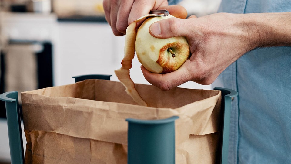 Närbild på händer och skalning av ett äpple men en lång skalremsa ovanför brun papperspåse för matavfall.