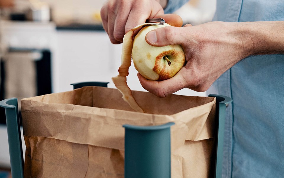 Närbild på händer och skalning av ett äpple men en lång skalremsa ovanför brun papperspåse för matavfall.